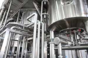Mirror-polished stainless steel tanks and intricate sanitary piping with valves, sight glasses, and support framework inside a modern dairy or food processing facility
