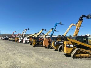 Row of heavy construction equipment including Caterpillar and Bobcat skid-steer loaders, telehandlers, and telescopic forklifts parked in an outdoor yard under a clear blue sky