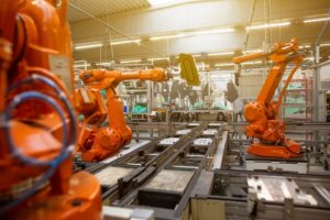 Orange industrial robotic arms performing automated assembly and material handling on a production line inside a modern, brightly lit manufacturing facility.