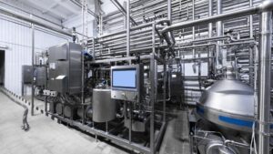Modern dairy or food processing plant interior with extensive sanitary stainless steel piping, automated valves, control panels, centrifugal pumps, and a large separator or clarifier in a clean, hygienic production hall.